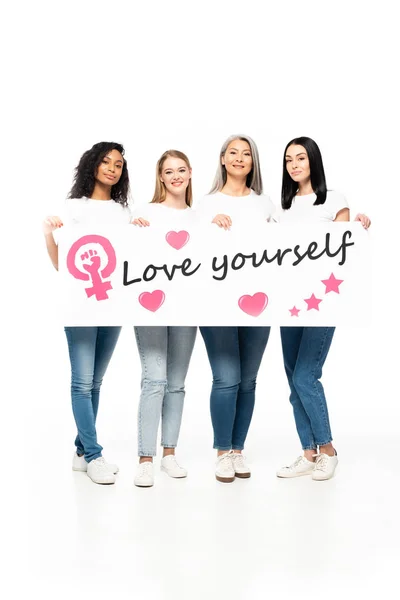 Happy multicultural women in denim jeans holding placard with love yourself lettering isolated on white — Stock Photo