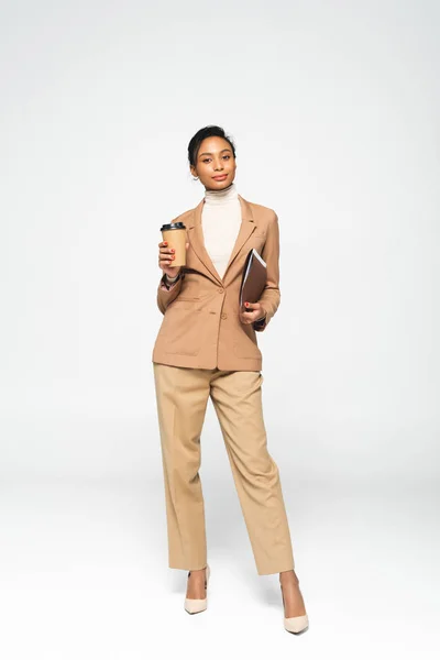 Atractiva mujer de negocios afroamericana sosteniendo carpeta y taza de papel en blanco - foto de stock