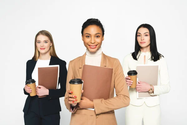 Happy multicultural businesswomen holding folders and paper cups isolated on white — Stock Photo