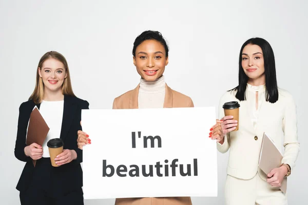 Cheerful multicultural businesswomen holding folders and paper cups near placard with i`m beautiful lettering isolated on white — Stock Photo