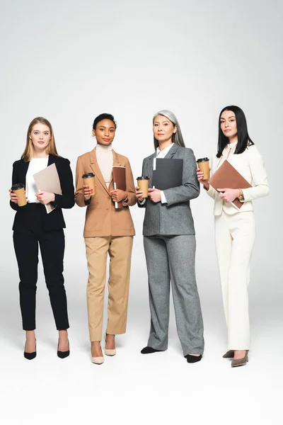 Vier multikulturelle Geschäftsfrauen halten Ordner und Pappbecher in der Hand, während sie auf weiß stehen — Stockfoto