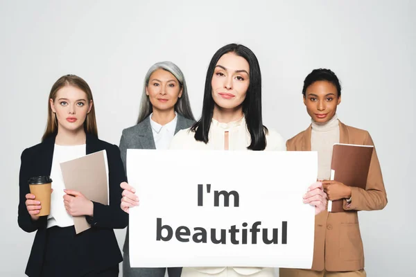 Multicultural businesswomen holding folders and disposable cups near placard with i`m beautiful lettering isolated on white — Stock Photo