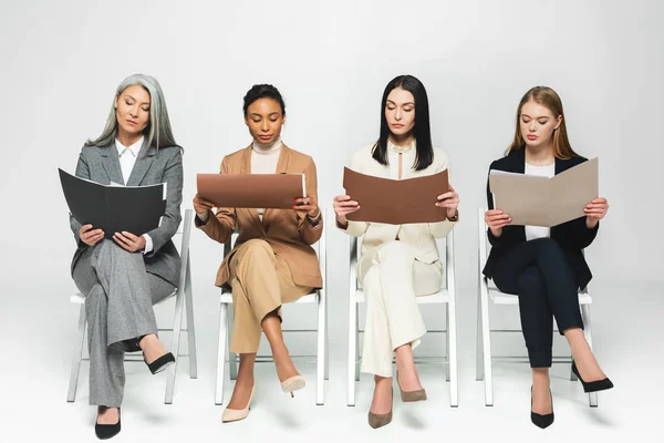 Four multicultural businesswomen in suits sitting on chairs and looking at folders on white — Stock Photo