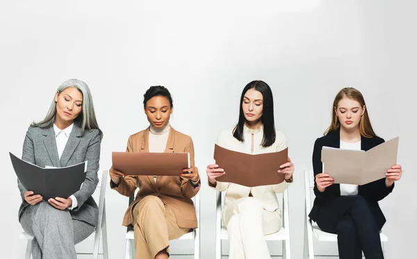 Four multicultural businesswomen in suits sitting on chairs and looking at folders isolated on white — Stock Photo