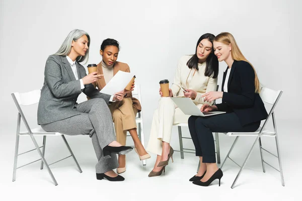 Fröhliche multikulturelle Geschäftsfrauen sitzen auf Stühlen mit Pappbechern und Laptop auf weiß — Stockfoto