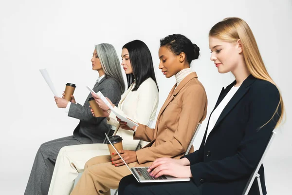 Side view of multicultural businesswomen sitting on chairs with paper cups and laptop on white — Stock Photo
