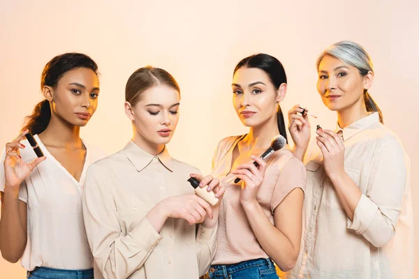 Cuatro mujeres multiculturales que sostienen cosméticos decorativos aislados en beige - foto de stock
