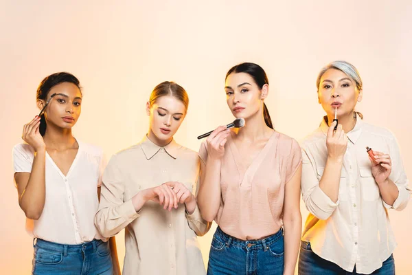 Atractivas mujeres multiculturales que sostienen cosméticos decorativos y cepillos cosméticos aislados en beige - foto de stock