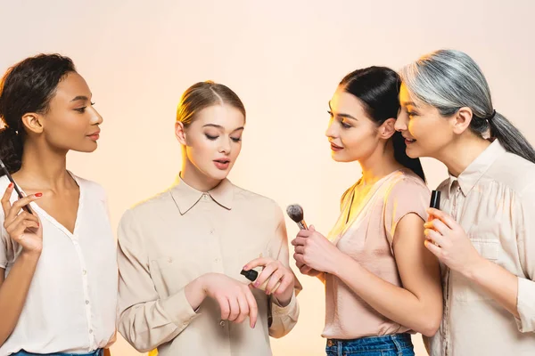 Attractive multicultural women holding decorative cosmetics isolated on beige — Stock Photo