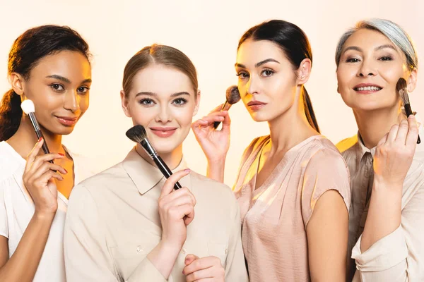 Beautiful multicultural women holding cosmetic brushes isolated on beige — Stock Photo