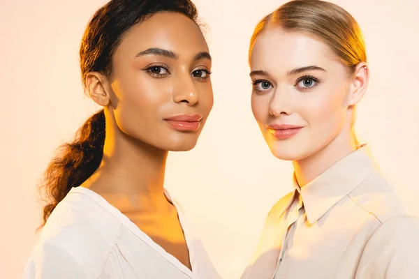 Two attractive multicultural women with makeup looking at camera isolated on beige — Stock Photo