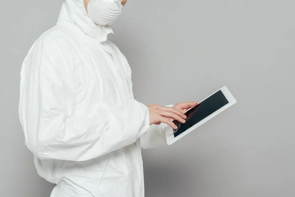 Vista recortada del epidemiólogo en traje de materiales peligrosos y máscara respiratoria que sostiene la tableta digital con pantalla en blanco sobre fondo gris - foto de stock