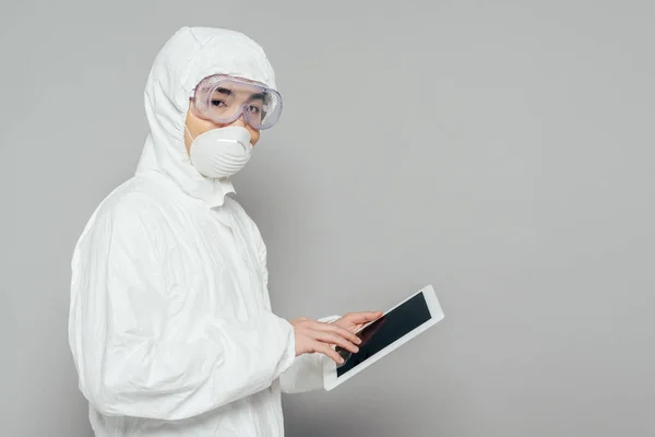 Азиатский эпидемиолог в защитном костюме и респираторной маске держит цифровой планшет с чистым экраном и смотрит на камеру на сером фоне — стоковое фото