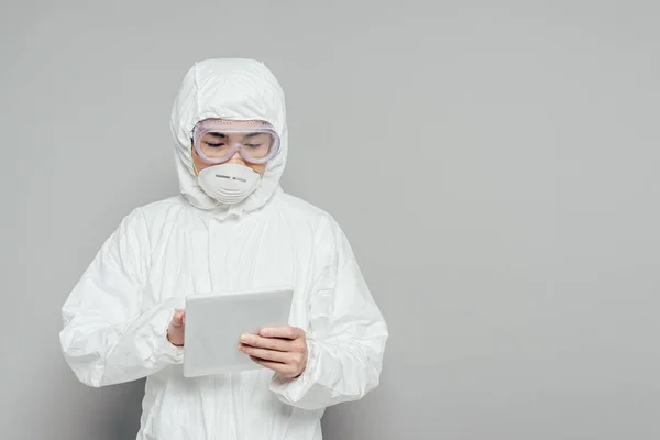 Epidemiólogo asiático en traje de hazmat y máscara respiratoria usando tableta digital sobre fondo gris - foto de stock