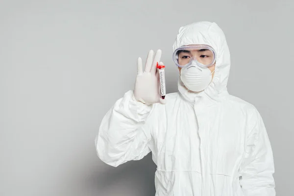 Epidemiólogo asiático en traje de hazmat y máscara respiratoria que muestra probeta con muestra de sangre sobre fondo gris - foto de stock