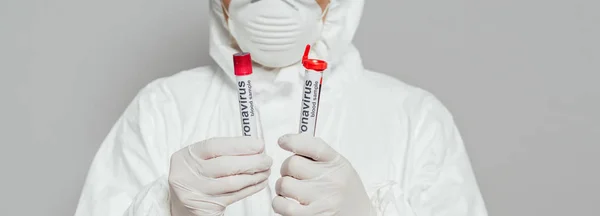 Cropped view of epidemiologist in hazmat suit and respirator mask holding test tubes with blood samples isolated on grey, panoramic shot — Stock Photo