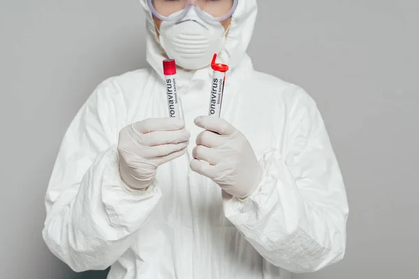 Vista recortada del epidemiólogo en traje de materiales peligrosos y máscara respiratoria que sostiene tubos de ensayo con muestras de sangre sobre fondo gris - foto de stock