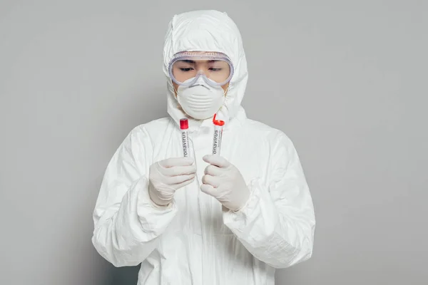 Asian epidemiologist in hazmat suit and respirator mask holding test tubes with blood samples on grey background — Stock Photo