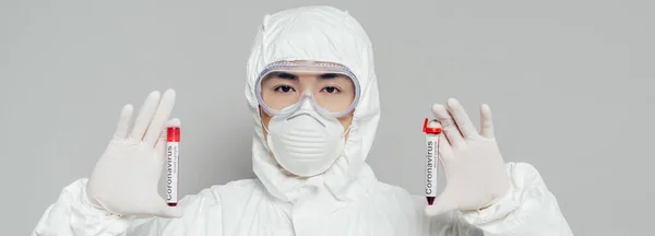 Panoramic shot of asian epidemiologist in hazmat suit and respirator mask showing test tubes with blood samples isolated on grey — Stock Photo