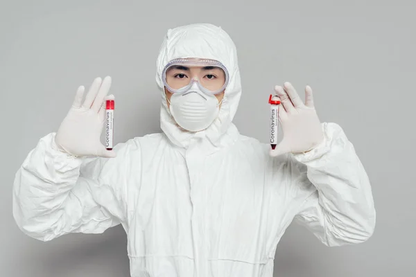 Épidémiologiste asiatique en combinaison Hazmat et masque respirateur montrant des éprouvettes avec des échantillons de sang sur fond gris — Photo de stock