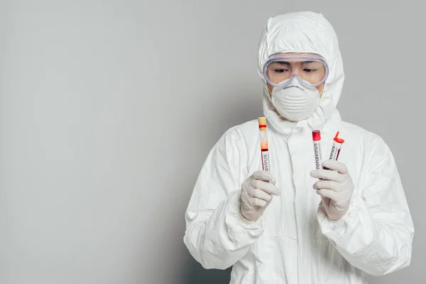 Asian epidemiologist in hazmat suit and respirator mask holding test tubes with blood samples on grey background — Stock Photo