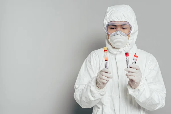 Asiatischer Epidemiologe in Warnanzug und Atemmaske mit Reagenzgläsern mit Blutproben auf grauem Hintergrund — Stockfoto