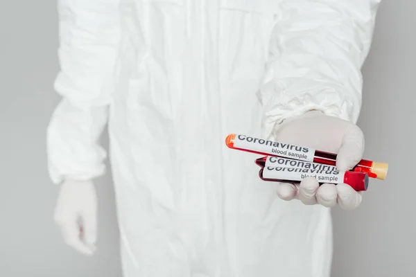 Partial view of epidemiologist in hazmat suit showing test tubes with blood samples on grey background — Stock Photo