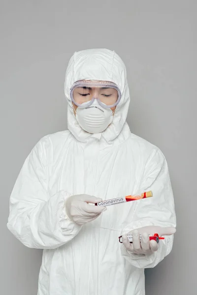 Epidemiólogo asiático en traje de hazmat y máscara respiratoria celebración de tubos de ensayo con muestras de sangre sobre fondo gris - foto de stock