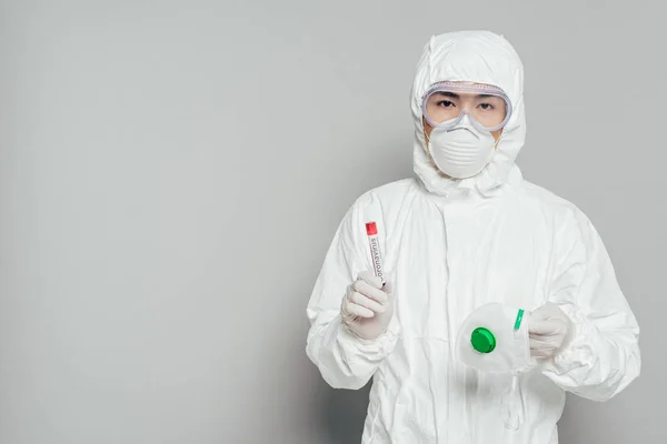 Азиатский эпидемиолог в защитном костюме, смотрящий в камеру, держа респираторную маску и пробирку с образцом крови на сером фоне — стоковое фото