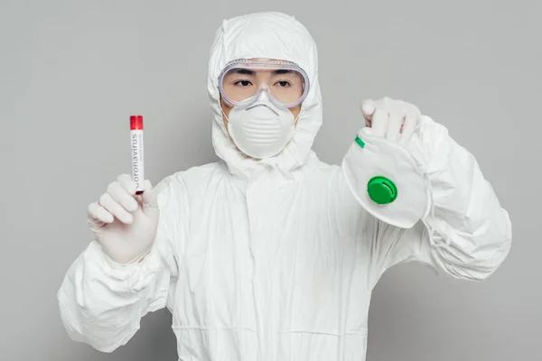 Epidemiólogo asiático en traje hazmat celebración de máscara respiratoria y probeta con muestra de sangre sobre fondo gris - foto de stock