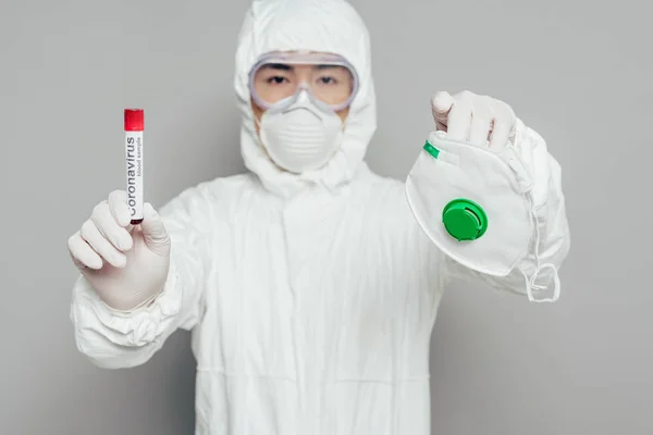 Asiatischer Epidemiologe im Warnanzug blickt in die Kamera, während er Atemschutzmaske und Reagenzglas mit Blutprobe auf grauem Hintergrund hält — Stockfoto
