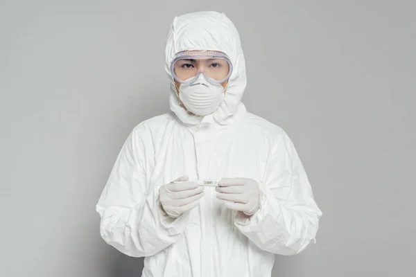 Épidémiologiste asiatique regardant la caméra tout en tenant thermomètre montrant une température élevée sur fond gris — Photo de stock