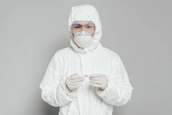 Asiatischer Epidemiologe blickt in die Kamera, während er das Thermometer hält, das hohe Temperaturen auf grauem Hintergrund zeigt — Stockfoto