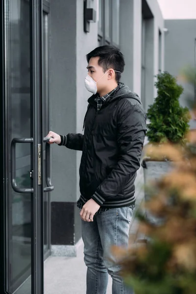 Selective focus of young asian man in respirator mask opening door and entering building — Stock Photo