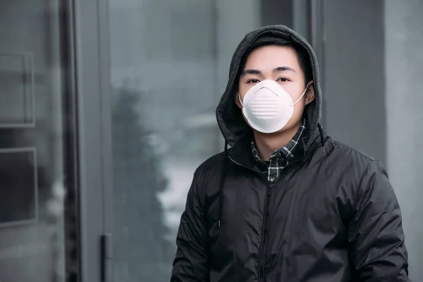 Giovane asiatico uomo in respiratore maschera guardando fotocamera mentre in piedi su strada — Foto stock