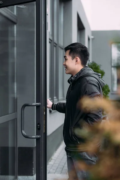 Selective focus of young asian man smiling while opening door and entering building — Stock Photo
