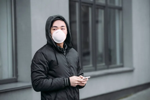 Young asian man in respirator mask holding smartphone and looking away while standing on street — Stock Photo