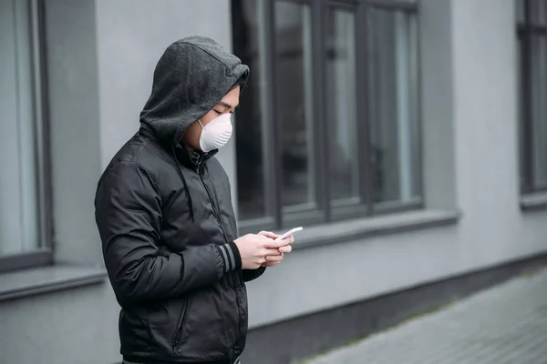 Молодий азіатський чоловік в респіраторній масці спілкується на смартфоні, стоячи на вулиці — стокове фото