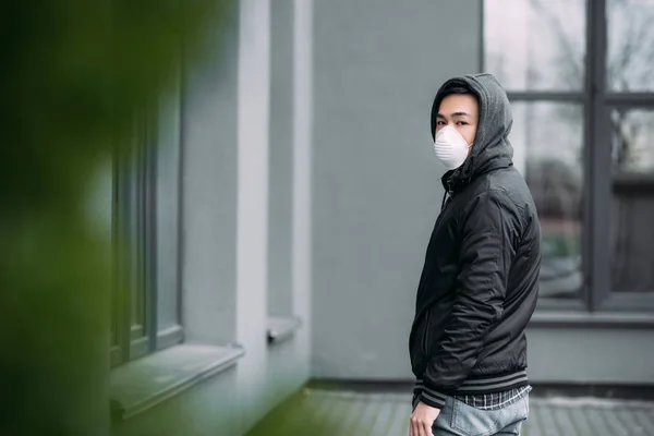 Enfoque selectivo de joven asiático hombre en respirador máscara mirando cámara mientras de pie en la calle - foto de stock