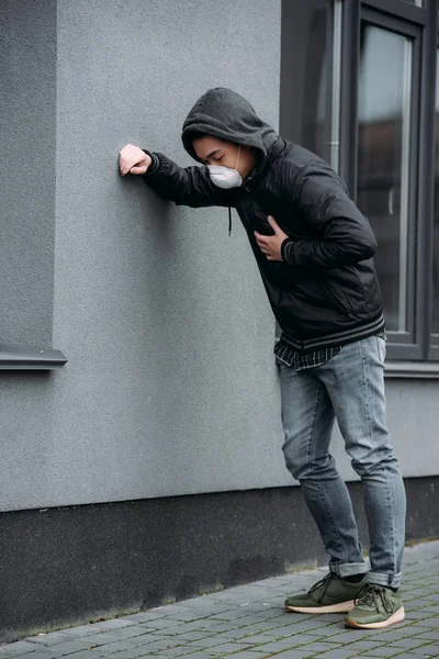 Giovane uomo in maschera respiratore appoggiato sulla parete e toccante petto mentre soffre di dolore polmonare — Foto stock