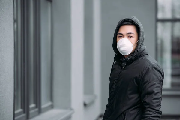 Joven asiático hombre en respirador máscara mirando cámara mientras de pie en calle - foto de stock