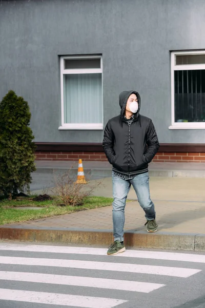 Joven asiático hombre en respirador máscara mirando lejos y cogido de la mano en bolsillos mientras cruzando camino - foto de stock