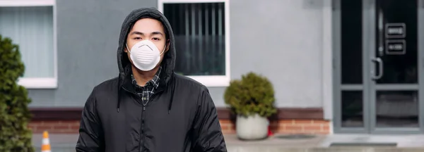 Panoramic shot of young asian man in respirator mask looking at camera while standing on street — Stock Photo