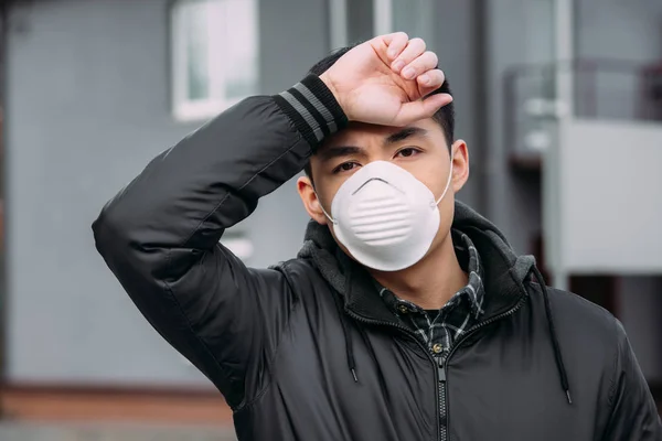 Joven asiático hombre en respirador máscara mirando cámara mientras padece de dolor de cabeza y tocando la frente - foto de stock