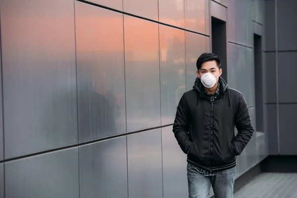 Joven asiático hombre en respirador máscara mirando cámara mientras caminando a lo largo de pared con manos en bolsillos - foto de stock