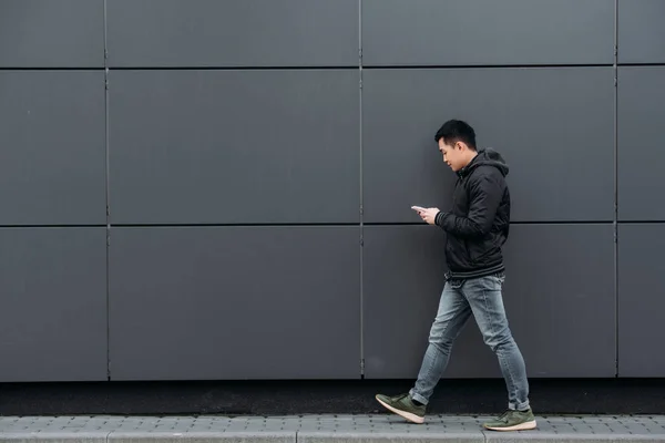 Seitenansicht eines jungen asiatischen Mannes, der auf dem Smartphone chattet, während er an der Wand entlang geht — Stockfoto