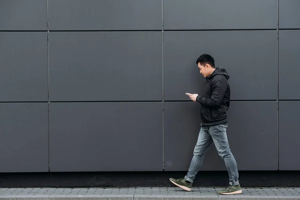 Side view of young asian man chatting on smartphone while walking along wall — Stock Photo
