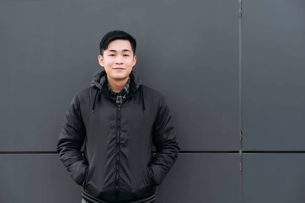 Jovem asiático homem sorrindo para câmera enquanto em pé por parede e segurando as mãos em bolsos — Fotografia de Stock