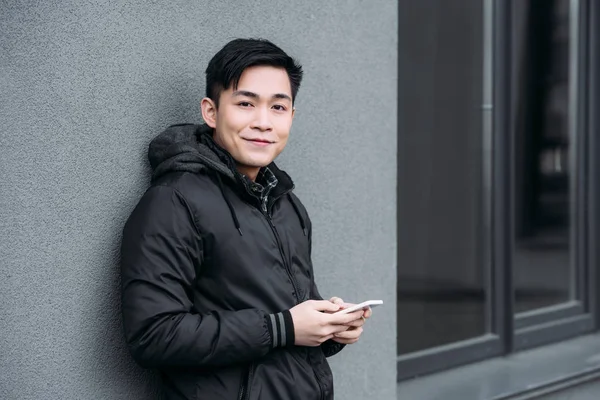 Young asian man smiling at camera while standing by grey wall and chatting on smartphone — Stock Photo