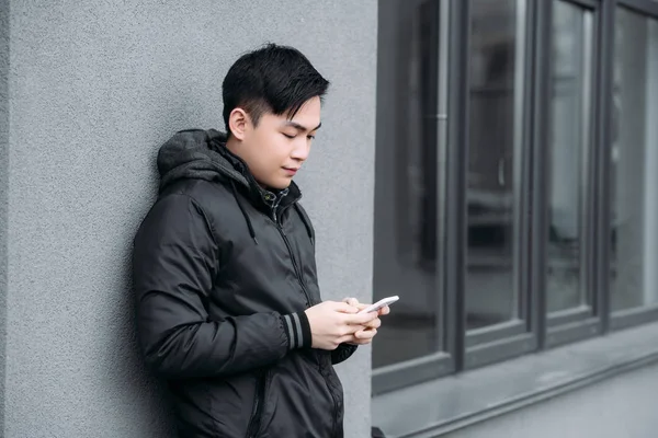 Young asian man chatting on smartphone while standing by grey wall — Stock Photo
